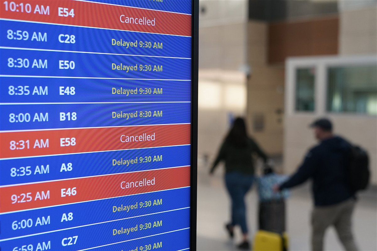 Travelers walk as a video board shows flight delays and cancellations at Ronald Reagan Washington National Airport in Arlington