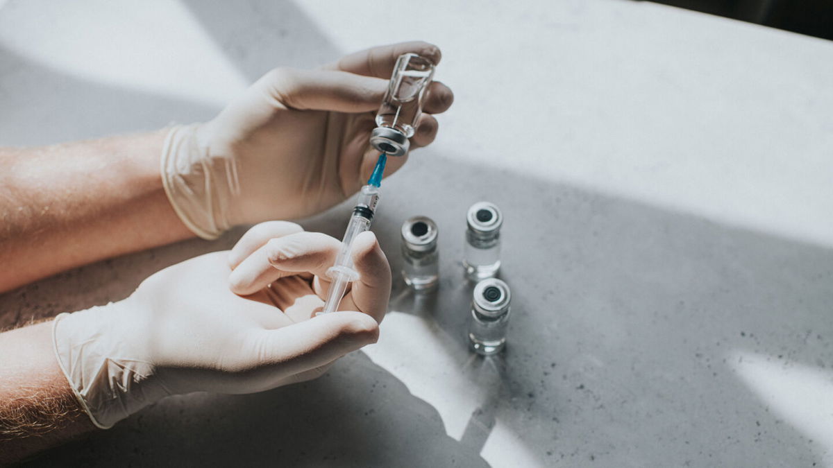 A hand wearing latex gloves hold a phial of clear liquid and a syringe. The medical professional uses the syringe to extract the medicine from the glass bottle. Conceptual with space for copy.