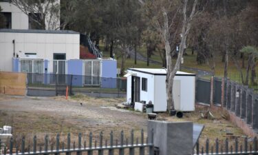 A portable security shed surrounded by weeds and discarded building materials on vacant land