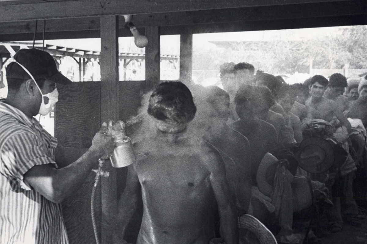 This photo taken in Hidalgo, Texas, in 1956 shows a masked worker spraying braceros with DDT while others wait in line.